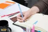 A person's hand is writing in a notebook with a blue pen. Other writing utensils and notebooks are on the table. Voir les images du produit