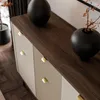 A close-up view of a modern dresser with gold knobs and a dark wood top, featuring two black vases as decor. Produktbild-Vorschau