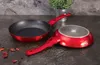 Two red frying pans are displayed on a dark countertop, with a potted plant in the background. Náhled obrázku produktu
