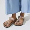 A close-up shot of a person's feet wearing copper Birkenstock Gizeh sandals, with light blue denim pants visible. Produktbild-Vorschau
