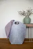 A grey felt laundry basket with a purple blanket draped over it, placed on a wooden floor next to a table with a green vase and branches. Produktbild-Vorschau