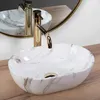 A close-up shot of a marble-patterned vessel sink with a gold faucet, placed on a wooden surface, with a mirror reflecting the sink and faucet in the background. Podgląd zdjęcia produktu