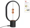A black oval-shaped desk lamp with two copper spheres suspended in the middle by a thin wire, sitting on a black cylindrical base. The lamp has an LED light strip around the inside of the oval frame. There are award logos on the right side of the image. Produktbild-Vorschau