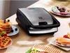 A multi-functional grill and waffle maker is shown on a wooden table, surrounded by various breakfast items like waffles, pancakes, donuts, and sandwiches. Voir les images du produit