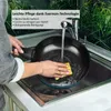 A person is washing a black frying pan in a kitchen sink under running water. Produktbild-Vorschau