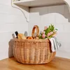 A wicker picnic basket filled with bread, fruit, and flowers sits on a wooden countertop, with a white tiled wall and shelf in the background. Produktbild-Vorschau