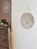 A round gold-framed mirror hangs on a textured white wall, reflecting a white dresser. To the left, a wooden dresser holds two dark green and gold vases, and a large pampas grass plume stands in the foreground. Produktbild-Vorschau