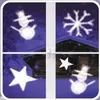 A window divided into four panes, each showing a projected winter-themed image: a snowman, a snowflake, and two stars. The background appears to be a snowy landscape with a wooden cabin. Produktbild-Vorschau