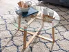 A round glass coffee table with a wooden base is placed on a patterned rug in a living room setting. Náhľad obrázku produktu