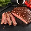 A close-up of sliced flank steak seasoned with salt and pepper, with cherry tomatoes and herbs in the background. Produktbild-Vorschau