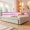 A modern bedroom with a beige upholstered daybed featuring integrated pink LED lighting. The bed has a pull-out trundle and is styled with pillows and a green throw blanket. A laptop is open on the bed. Produktbild-Vorschau
