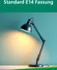 A black desk lamp with its light on, casting a warm glow on a teal background. Produktbild-Vorschau