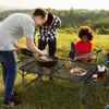 People are having a barbecue outdoors on a sunny day, with a folding table set up for food. Produktbild-Vorschau