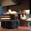 A projector is placed on a wooden table in a modern living room setting, with a woman working on a laptop in the background. Anteprima dell’immagine prodotto