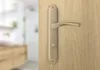 A close-up of a bronze door handle on a wooden door, with a blurred interior in the background. Produktbild-Vorschau