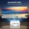 A white mini projector is shown in the foreground, projecting a vibrant image of a sunset beach scene onto a large screen in the background. The projector is placed on a reflective surface. Produktbild-Vorschau