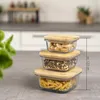 Three glass food storage containers with bamboo lids are stacked on a countertop, with a blurred kitchen background and a plant in the foreground. Produktbild-Vorschau