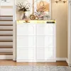 A white shoe cabinet with six flip-down doors and an open shelf with LED lighting, placed in a hallway next to a staircase. Decorative items are on top of the cabinet. Náhľad obrázku produktu