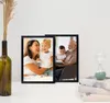 Two black picture frames are displayed on a white surface, with a vase of dried reeds to the left and a stack of books with an hourglass to the right. Produktbild-Vorschau
