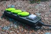 A black power strip with three green outlets and a white switch is lying on the ground, which is covered with small stones and dirt. There are water droplets on the power strip, suggesting it is outdoors and possibly raining. Produktbild-Vorschau