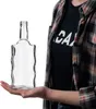 A person is holding a clear glass bottle with a unique wavy design on a white background. Produktbild-Vorschau