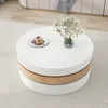 A round coffee table with a slice of cake and a vase of flowers on top, placed on a rug in a living room setting. Produktbild-Vorschau