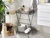 A bathroom setting with a black metal side table holding towels and decorative items, next to a woven basket and a sink. Produktbild-Vorschau