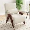 A cream-colored armchair with wooden legs and armrests, placed on a patterned rug in a well-lit room with a large window and a plant. Náhled obrázku produktu