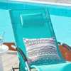 A close-up of a turquoise lounge chair with a patterned pillow and a glass of water next to it, with a swimming pool in the background. Náhled obrázku produktu