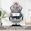 A man is sitting cross-legged in a gaming chair with a laptop on his lap. A plant is visible to the left, and a floor lamp to the right. Produktbild-Vorschau