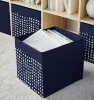 A dark blue storage cube with a white polka dot pattern sits on a light gray rug in front of a bookshelf. Produktbild-Vorschau