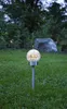 A solar-powered garden light with a cracked glass globe sits in a grassy yard at dusk. Produktbild-Vorschau