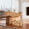 A cardboard box filled with wood briquettes sits on a wooden floor in a living room setting, with some briquettes spilled out in front of the box. The background shows a blurred view of a city skyline through a large window and a fireplace. Anteprima dell’immagine prodotto