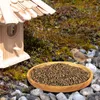 A bird is eating from a bowl of birdseed next to a wooden birdhouse in a garden setting. Produktbild-Vorschau