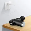 A black massage gun is charging on a wooden shelf, plugged into a white wall socket. Produktbild-Vorschau