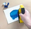 A hand is holding a yellow and black encaustic iron, applying blue paint to a white card. Several blue wax blocks are visible in the upper left corner. Náhľad obrázku produktu