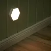 A hexagonal LED light is mounted on a paneled wall above a white baseboard, casting a warm glow on the wall and the wooden floor. Produktbild-Vorschau