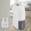 A white dehumidifier is shown in a bedroom setting, next to a wall-mounted electrical outlet with a digital display. Podgląd zdjęcia produktu
