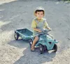 A young child is sitting on a toy tractor with a trailer attached, outdoors on a dirt path. Produktbild-Vorschau