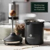 A close-up shot of a manual milk frother and a milk frothing pitcher, with a coffee grinder and jars of coffee beans in the background. Produktbild-Vorschau