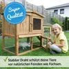 A young girl is sitting next to a wooden rabbit hutch in a grassy backyard. Produktbild-Vorschau