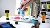 A hand is squeezing a tube of blue acrylic paint onto a palette with other colors, with several tubes of paint in the background. Anteprima dell’immagine prodotto