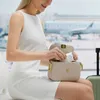 A woman is at an airport, taking a small white item out of her beige handbag. An airplane is visible through the window in the background. Náhled obrázku produktu