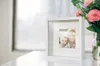 A white shadow box picture frame with a photo of a woman and a baby inside, placed on a white surface next to a vase of pink flowers. Produktbild-Vorschau