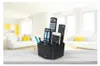 A black remote control organizer filled with remotes, a smartphone, pens, and glasses, placed on a wooden table in front of a blurred living room background. Náhľad obrázku produktu