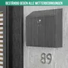 A black mailbox with a key in the lock, mounted on a concrete wall during a rain shower. The house number '89' is visible below the mailbox. Produktbild-Vorschau