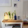 A bedside table with a lamp, flowers in vases, and framed pictures. Produktbild-Vorschau
