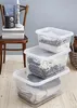 Three clear plastic storage boxes are stacked on a wooden floor, filled with folded textiles. A wooden chair with books and a radio is in the background. Voir les images du produit