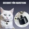 A white cat wearing a collar with a bell and a black charm, with a close-up inset of the charm. Produktbild-Vorschau