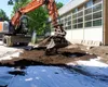 An excavator is digging and moving soil on a construction site next to a building. Produktbild-Vorschau